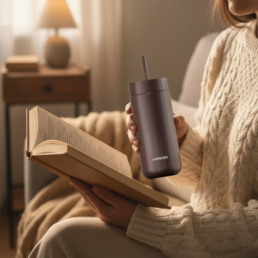 Person holding brown Lepresso tumbler with straw while reading a book indoors